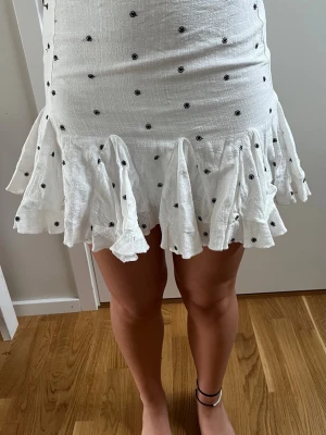Vit kortkjol med volang och prickar - Supersöt vit kortkjol med svarta prickar och volangkant. Kjolen har en figurnära passform upptill och faller ut i en volangig nederdel som ger en lekfull vibe. Perfekt för varma dagar och somriga outfits.