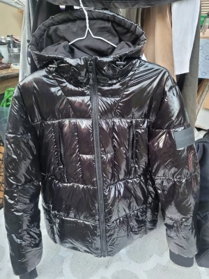 Svart glansig pufferjacka Calvin Klein - Snygg svart pufferjacka från Calvin Klein med glansigt material och stor huva. Jackan har dragkedja framtill, två sidofickor och CK-logga broderad på huvan. Perfekt för kalla vinterdagar och ger en modern look.storlek S men som en M