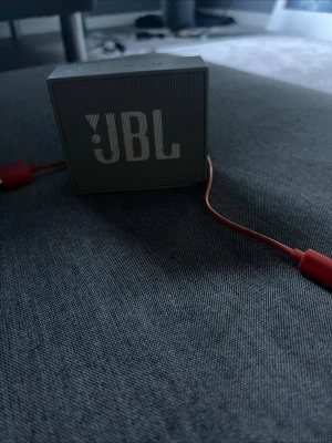 JBL trådlös högtalare grå - Smidig och portabel trådlös högtalare från JBL i grå färg. Kubformad design med tydlig JBL-logga på framsidan. Tillverkad i plast och metallgaller, med medföljande röd USB-laddkabel. Perfekt för att ta med sig överallt och spela musik trådlöst.