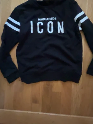Svart sweatshirt Dsquared2 ICON - Svart sweatshirt från Dsquared2 med vit text 'ICON' och logga på bröstet. Tröjan har vita dubbla ränder på ärmarna och rund hals. Perfekt för en streetwear-look och enkel att matcha med jeans eller joggers.