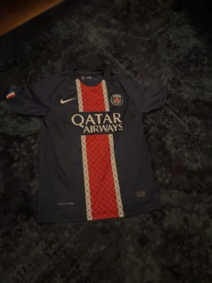 PSG matchtröja med Qatar Airways - Mörkblå PSG matchtröja från Nike med röd och vit rand i mitten och Qatar Airways-logga på bröstet. Tröjan har klubbmärke på vänster sida och Nike-logga på höger. Korta ärmar och sportig passform, perfekt för fotboll eller träning.