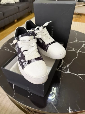 Numeris skor  - Snygga svarta sneakers med vit splattermönster och breda vita skosnören. Skorna har vit tå och sula i gummi samt låg profil. Perfekta för dig som vill sticka ut med en unik och trendig look.
