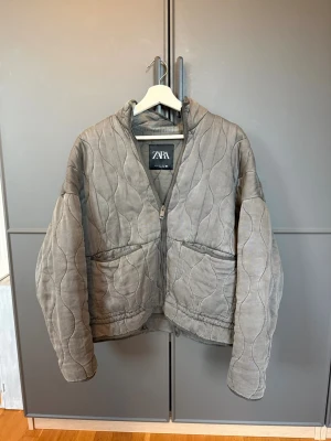 Grå quiltad pufferjacka från Zara - Snygg grå pufferjacka från Zara med quiltat mönster och hög krage. Jackan har dragkedja framtill och två stora fickor. Tillverkad i mjuk manchester som ger en unik look. Perfekt för dig som vill ha en trendig och bekväm jacka till höst och vinter.
