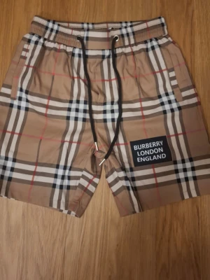 Rutig Burberry shorts med snörning - Snygga shorts från Burberry med klassiskt rutigt mönster i beige, svart, vitt och rött. Shortsen har elastisk midja med svart snörning och en patch med texten 'Burberry London England' på ena benet. Perfekta för sommaren och ger en trendig look.