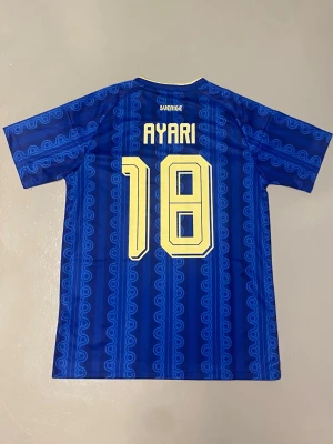 VM 2026 Sverige fotbollströja Adidas ayari#18 - Blå fotbollströja från Adidas med Sveriges landslagsemblem och nummer 18 i gult. Tröjan har korta ärmar, gul krage och tre gula ränder på axlarna. Namnet 'Ayari' står på ryggen. Tillverkad i lätt och ventilerande material.