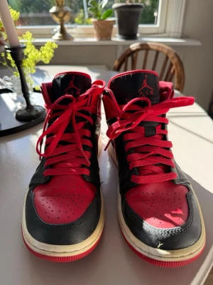 Air Jordan 1 svart/röd sneakers - Säljer ett par klassiska Air Jordan 1 sneakers i svart och rött läder med röda skosnören och vit sula. Ikonisk Nike Swoosh på sidan och Air Jordan-logga vid ankeln. Perfekta för dig som gillar streetwear och vill sticka ut.