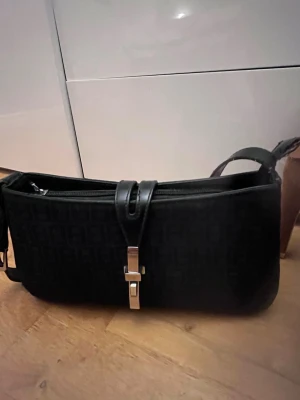 Svart axelremsväska med metallspänne - Snygg svart handväska med axelrem och ett stilrent metallspänne framtill. Väskan har en avlång form och diskret mönster i materialet. Perfekt för dig som gillar minimalistisk och trendig design. Dragkedja upptill för extra säkerhet. Lite smutsig på insidan man går att tvätta