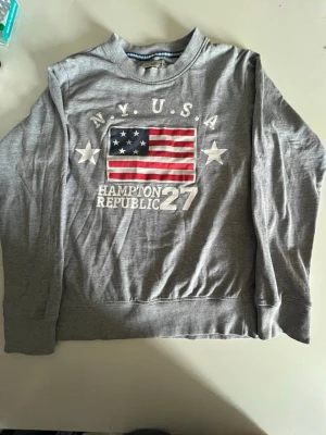 Grå sweatshirt med USA-flagga  - Snygg grå sweatshirt med tryck av amerikanska flaggan och texten 'N.Y. U.S.A Hampton Republic 27' framtill. Tröjan har rund halsringning och långa ärmar. Perfekt för en avslappnad och sportig stil.