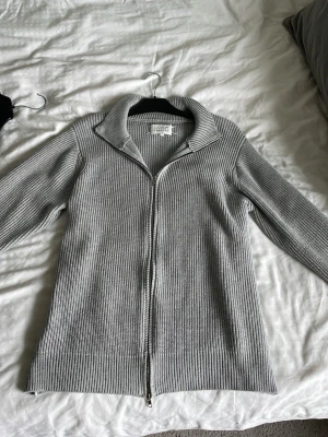Grå stickad ziptröja Maison Margiela - Säljer en stilren grå stickad ziptröja från Maison Margiela. Tröjan har ribbstickad struktur, hög krage och dragkedja framtill. Perfekt för lager på lager och ger en clean look till din outfit.