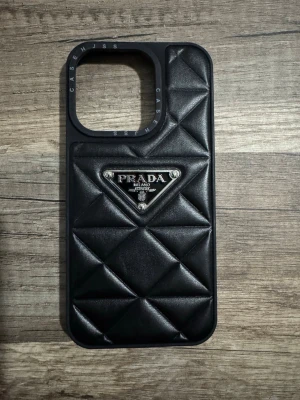 Mobilskal iPhone 15 Pro Prada - Snyggt svart mobilskal med quiltad design och metallogo från Prada. Skalet har en lyxig känsla med mjukt, vadderat material och passar perfekt för dig som vill ha en exklusiv look på mobilen.