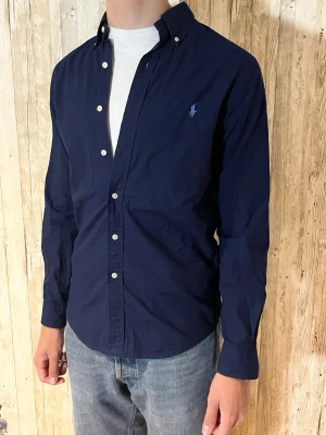 Mörkblå skjorta med knappar - Stilren mörkblå skjorta med klassisk krage och vita knappar. Skjortan har lång ärm och en broderad logga på bröstet. Perfekt för dig som gillar en enkel men snygg look. Tillverkad i bomull för en skön känsla.