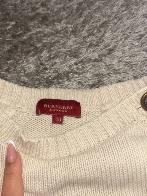 Krämvit stickad tröja Burberry - Snygg krämvit stickad tröja från Burberry med broderad logga framtill och tre dekorativa knappar på axeln. Tröjan har ribbade muddar och är tillverkad i mjukt stickat material. Perfekt för dig som gillar klassisk och stilren design. (Orginalpris är 15 689kr då den är vintage)