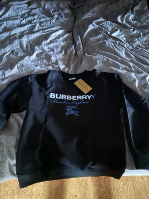 Svart Burberry sweatshirt - Snygg svart sweatshirt från Burberry med blått tryck framtill där det står 'Burberry London, England' och en ikonisk riddare. Tröjan har rund halsringning och ribbade muddar vid ärmslut och nederkant. Perfekt för en trendig och avslappnad stil.