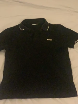 Svart pikétröja från BOSS - Klassisk svart pikétröja från BOSS med vit logga på bröstet och vita detaljer på kragen och ärmsluten. Tröjan har korta ärmar och en mjuk krage med knappar framtill. Perfekt för en clean och stilren look.