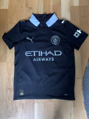 Manchester City svart matchtröja Puma - Säljer en svart Manchester City matchtröja från Puma med vit krage och tryck i vitt. På bröstet finns klubbmärke och sponsorlogga, på ryggen står det Foden och nummer 47. Perfekt för fotbollsträning eller att bära som supporter.