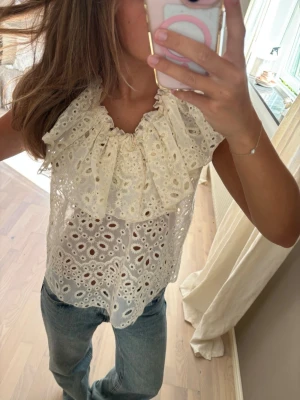 Vit ärmlös blus med volang - Säljer en superfin vit blus med broderade hålmönster och stor volang runt halsen. Blusen är ärmlös och har en romantisk, luftig känsla som passar perfekt till jeans eller kjol. Perfekt för dig som gillar detaljer och en feminin stil.