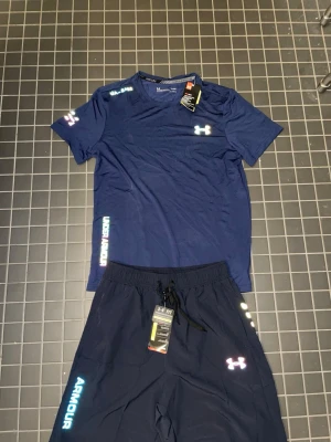 Blått träningsset från Under Armour - Snyggt träningsset med t-shirt och shorts från Under Armour. Båda plaggen är mörkblå med vita och blå logotyper. T-shirten har rund hals och korta ärmar, shortsen har dragsko i midjan och tryckt logga på benet. Perfekt för gym eller löpning.