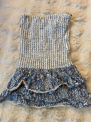 Vit och blå bandeauklänning Gina Tricot - Supersöt bandeauklänning från Gina Tricot i vitt och blått med smock upptill och volangig kjol. Klänningen har ett unikt mönster och är perfekt för varma dagar. Tillverkad i mjuk bomull som känns skön mot huden.