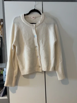 Vit kofta från Gina Tricot - Passar även XS/S. Mysig vit kofta från Gina Tricot med rund halsringning och knappar framtill. Koftan har ribbade muddar vid ärmslut och nederkant, och är tillverkad i ett mjukt stickat material. Perfekt att slänga på sig när det är kyligt ute.