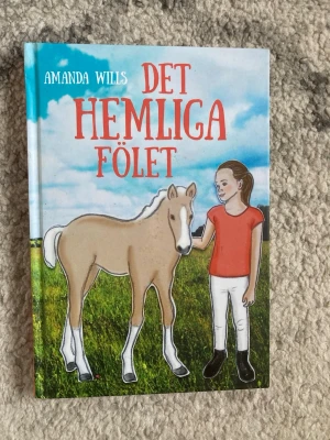 Det hemliga fölet av Amanda Wills - En färgglad bok med titeln 'Det hemliga fölet' av Amanda Wills. Omslaget visar en flicka i röd tröja och vita byxor bredvid ett ljust föl på en grön äng under blå himmel. Perfekt för dig som gillar hästar och spännande berättelser.