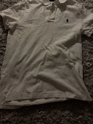 Vit pikétröja med krage - Klassisk vit pikétröja med krage och knappar framtill. Tröjan har korta ärmar och en broderad logotyp på bröstet. Perfekt för en clean och stilren look. Inga defekter alls på tröjan, skulle säga att storköpen motsvarar xs/s