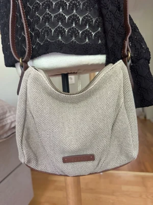 Beige ESPRIT axelväska i tyg med bruna detaljer - Snygg beige axelväska från ESPRIT i grovt tyg med bruna läderdetaljer och justerbar axelrem. Väskan har en mjuk, rundad form och stängs med dragkedja. Perfekt storlek för det viktigaste och enkel att matcha till olika outfits.
