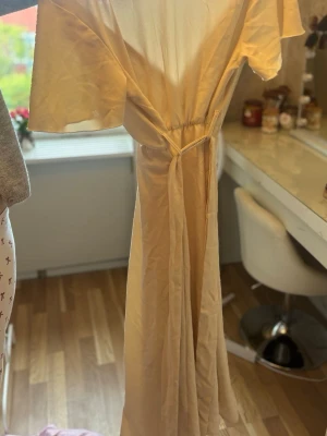 Lång beige omlottklänning med knyt - Säljer en elegant långklänning i beige med omlottmodell och knyt i midjan. Klänningen har korta ärmar och ett luftigt, lätt material som passar perfekt till varmare dagar. Enkel men stilren design som ger en avslappnad men chic look.har dock ett hål på armen.tror de är o storlek M men är inte säker 