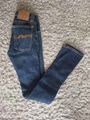 Mörkblå skinny jeans från Nudie - Mörkblå skinny jeans från Nudie med klassisk femficksdesign och orange kontrastsömmar. Jeansen har en smal passform och är gjorda i stretchigt denim för extra bekvämlighet. Snygg detalj med Nudie-loggan broderad på bakfickan.