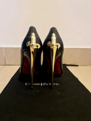 Svarta heels med gulddetaljer - Unika svarta pumps från Cesare Paciotti med höga, smala klackar i guld och röda sulor med spetsmönster. Skorna har en elegant siluett och exklusiva gulddetaljer bak på hälen som ger en lyxig känsla. Perfekta för dig som vill sticka ut.