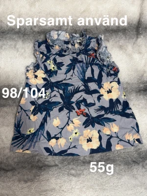 Blommig ärmlös topp med volang - Söt blommig topp i blå och beige toner med volangdetaljer vid hals och ärmhål. Ärmlös modell med rund halsringning. Perfekt för varma dagar och ger en fräsch look.🌸 Sparsamt använd. Storlek 98/104