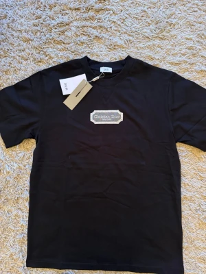 Svart t-shirt  - Svart t-shirt med rund hals och korta ärmar . På bröstet sitter en broderad beige patch T-shirten har en klassisk passform och är tillverkad i mjukt bomullstyg.