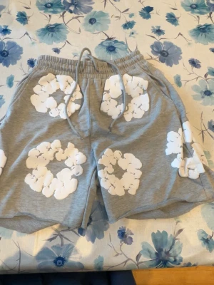 Grå shorts med vita blommor - Säljer ett par grå shorts med vita blommönster och snörning i midjan. Shortsen har avslappnad passform och elastisk midja för extra komfort. Perfekta för varma dagar och chill häng. Shortsen är i storlek S/////// Pris kan diskuteras