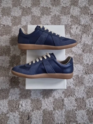 Maison margiela gats - Snygga marinblå sneakers med beige snörning och klassisk gummisula. Skorna har en mix av mocka och läderdetaljer som ger en stilren look. Perfekta för dig som vill ha en clean och tidlös sneaker till vardagsoutfiten. Liten defekt, syns lite på utsidan av skon också men inget man tänker på. 