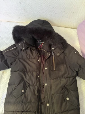 Svart dunjacka med fuskpäls - Snygg svart dunjacka med huva och fuskpälsdetalj. Jackan har dragkedja och knappar framtill, två snedställda fickor med lock samt en bröstficka med dragkedja. Perfekt för kalla vinterdagar och har en klassisk, puffig passform.
