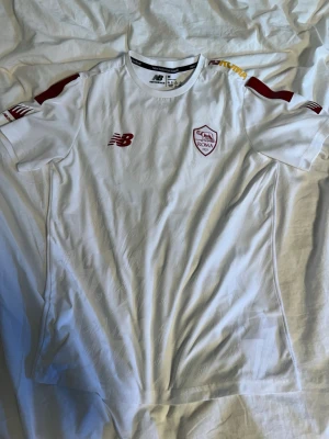 Vit AS Roma matchtröja New Balance - Snygg vit AS Roma matchtröja från New Balance med klubbmärke på bröstet och röda detaljer på axlar och logga. Tillverkad i lätt och ventilerande material, perfekt för träning eller match. Kortärmad och sportig passform.