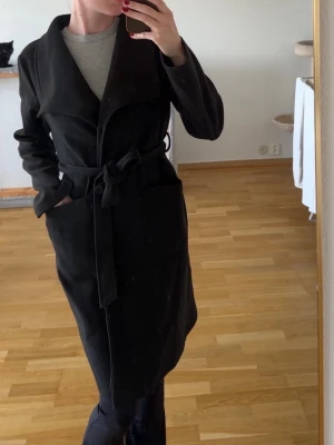 Svart kappa med knytskärp - Stilren svart kappa med bred krage och knytskärp i midjan. Kappan har stora fickor framtill och en rak passform som ger en snygg siluett. Perfekt för kyligare dagar och enkel att matcha med olika outfits.