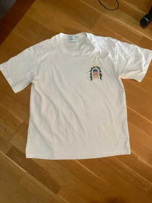 Vit t-shirt från Casablanca - Säljer en vit t-shirt från Casablanca Tennis Club i storlek XL. T-shirten har ett litet färgglatt tryck på bröstet och är gjord i mjuk bomull. Klassisk passform och rund hals, perfekt för en clean och stilren look.