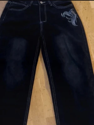 Mörkblå jeans med vit brodyr - Snygga mörkblå jeans med raka ben och vit kontrastsöm. På ena fickan finns en cool vit brodyr i form av ett abstrakt mönster. Jeansen har klassisk femficksmodell och är gjorda i denim. Perfekta för dig som vill sticka ut lite extra.Priset går att diskuteras vid snabb äffar 😊