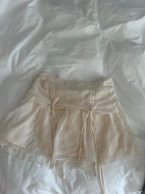 Beige kortkjol med knyt och volang - Söt beige kortkjol i ett tunt, luftigt material med dubbla lager och volangkant. Kjolen har dekorativa knytband framtill och bakom❤️otroligt fin
