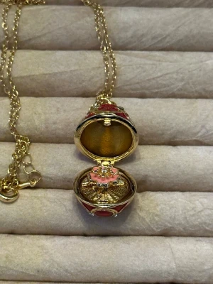Guldfärgat ägghalsband - Unikt halsband med ett guldfärgat hänge i form av ett ägg som går att öppna. Utsidan är dekorerad med röd emalj, små vita pärlor och guldfärgade detaljer. Inuti finns en liten rosa blomma. Kedjan är tunn och guldfärgad.
