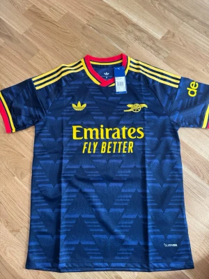 Mörkblå Arsenal tröja Adidas - Snygg mörkblå Arsenal matchtröja från Adidas med gula detaljer och röd krage. Tröjan har gula ränder på axlarna, tryckt Arsenal-logga och Emirates Fly Better på bröstet. Tillverkad i lätt och ventilerande material, perfekt för träning eller match.