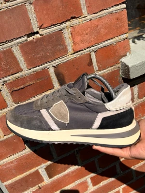 Philippe model sneakers - Snygga sneakers i olika grå nyanser med detaljer i mocka och mesh. Skon har en vit och svart sula med retrovibbar och en sköldliknande patch på sidan. Perfekt för dig som gillar stilrena och sportiga skor med en modern touch.