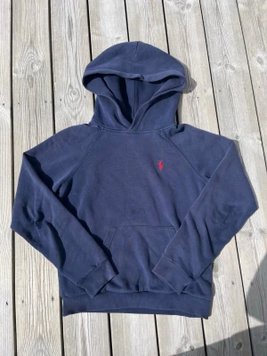 Mörkblå hoodie från Polo Ralph Lauren - Snygg mörkblå hoodie från Polo Ralph Lauren med klassisk röd logga broderad på bröstet. Hoodien har huva, känguruficka och ribbade muddar vid ärmslut och midja. Perfekt för en chill och avslappnad stil.