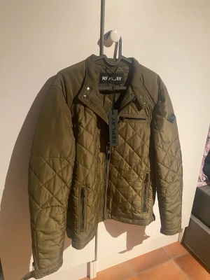 Olivgrön quiltad jacka från Replay - Snygg olivgrön quiltad jacka från Replay med dragkedja framtill, tre fickor med dragkedja och ståkrage med tryckknappar. Jackan har ett rutmönstrat yttertyg och Replay-logga på ärmen. Perfekt för dig som vill ha en stilren och trendig look. Köpt från pondus för 1999kr, skick 10/10. Hör av dig vid frågor:)