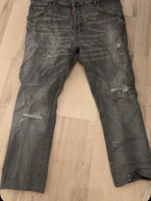 Dsquared2 Jeans - Säljer ett par grå jeans från Dsquared2 med distressed detaljer och slitningar på både fram- och baksida. Jeansen har raka ben, klassiska fem fickor och coola färgstänk för en edgy look. Perfekta för dig som gillar streetstyle och vill sticka ut. Det står 52 men passar 48. Vikta nere då de är lite kortare.