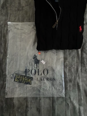 Svart stickad tröja Polo Ralph Lauren - Svart stickad tröja från Polo Ralph Lauren med halv dragkedja och röd broderad logga på bröstet. Tröjan har kabelstickat mönster, ribbade muddar och hög krage. Perfekt för dig som gillar klassisk och stilren design.