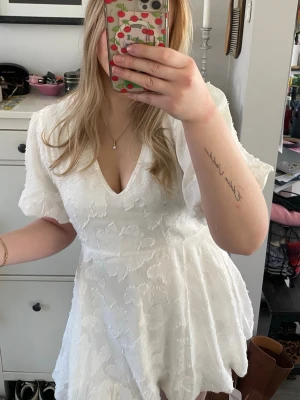 Studentklänning  - Söt vit kortklänning från Boohoo med v-ringning och korta puffärmar. Klänningen har ett diskret strukturmönster i tyget och en lätt utsvängd kjol. Perfekt för sommarens alla tillfällen!