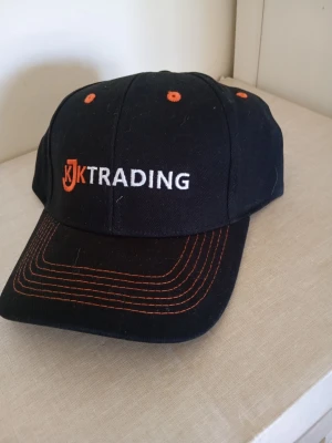 Svart keps med JK Trading-logga - Snygg svart keps från JK Trading med broderad logga i vitt och orange framtill. Kepsen har böjd skärm med orangea sömmar och ventilationshål i matchande färg. Perfekt för en sportig och avslappnad stil.