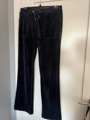 Mörkblå mjukisbyxor från Juicy Couture - Mysiga mörk blåa mjukisbyxor från Juicy Couture i velour med raka ben, dragsko i midjan och stora fickor både fram och bak. Klassisk logga broderad på ena fickan framtill. Perfekta för en chill och avslappnad stil. Dem är nedsydda men det går enkelt att ta upp styngen igen  utan problem eller tecken på att dem varit nedsydda om man vill det