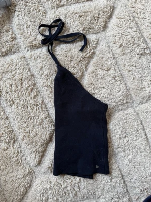 Svart  halterneck topp - Trendig svart oneshoulder topp med halterneck och knyt i nacken. Toppens material känns mjukt och stretchigt, perfekt för en cool och avslappnad stil. Enkel och stilren design utan mönster eller extra detaljer.